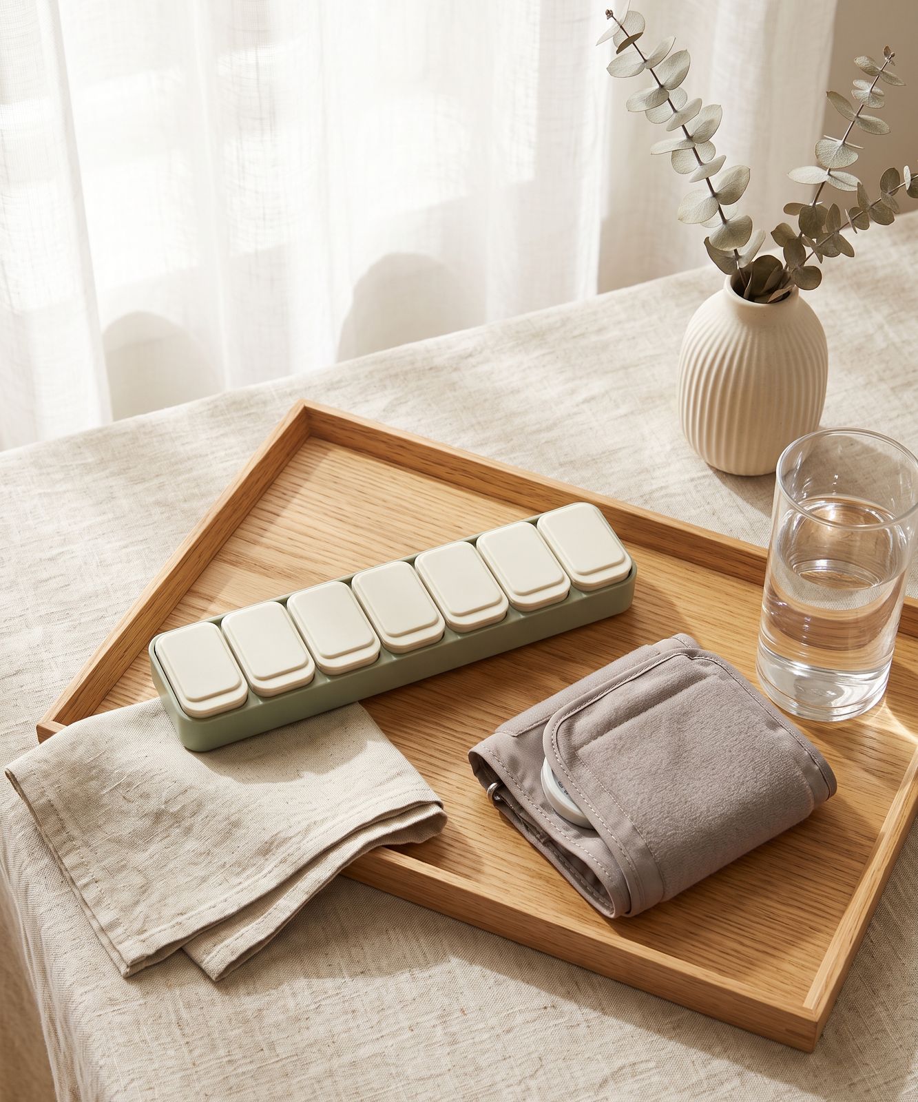Weekly medication organizer and blood pressure cuff arranged on a wooden tray with eucalyptus